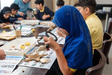 A girl in a blue hijab using a magnifying glass to examine a shell during an educational workshop on marine life.