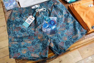 A pair of blubeach shorts patterned with outlines of turtles, seahorses, shells, starfish and types of fish. The label reads "Fatface X Marine Conservation Society". On top of the shorts is a leaflet that reads "Living without single-use plastic"
