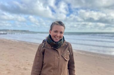 Trustee Debs Mahon smiles on a beach with the sea to the right side of her. She is wearing a brown jacket and has her hair braided.