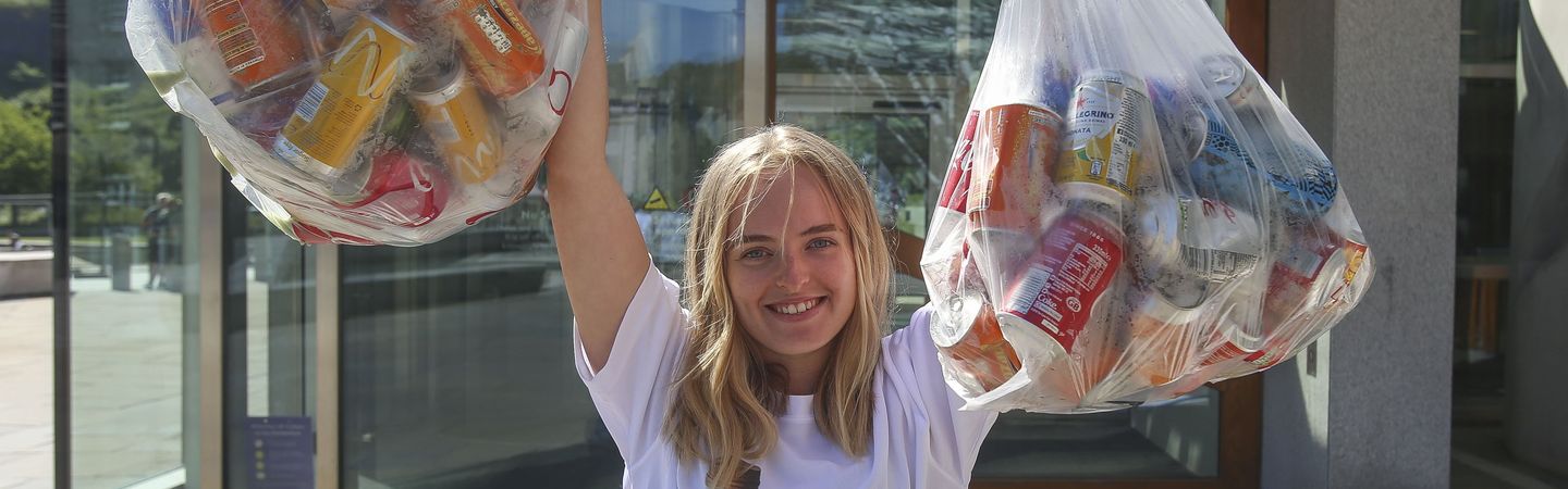 A female stands facing the camera wearing a white t-shirt that says "Have you got the bottle?". She has her arms raised and she is holding two bags filled with plastic bottles