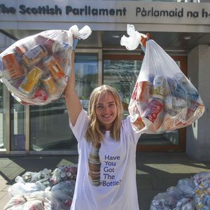 A female stands facing the camera wearing a white t-shirt that says "Have you got the bottle?". She has her arms raised and she is holding two bags filled with plastic bottles