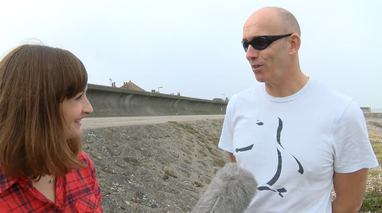 A man is being interviewed by a female presenter on a beach. They are near a concrete sea wall. The microphone can be seen at the bottom of the image.