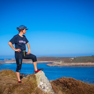 A woman with one leg resting upon a rock. She is overlooking a body of water with her hands on her hips. She is wearing a cap and a Marine Conservation Society t-shirt.