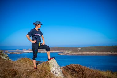 A woman with one leg resting upon a rock. She is overlooking a body of water with her hands on her hips. She is wearing a cap and a Marine Conservation Society t-shirt.