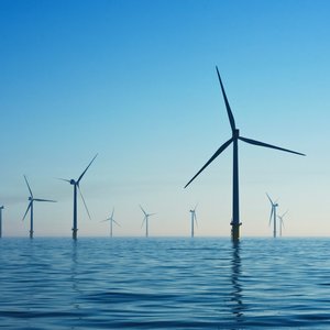 A wind farm out at sea shows around 6 turbines in the foreground with more in the far distance under a bright blue sky.