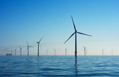 A wind farm out at sea shows around 6 turbines in the foreground with more in the far distance under a bright blue sky.