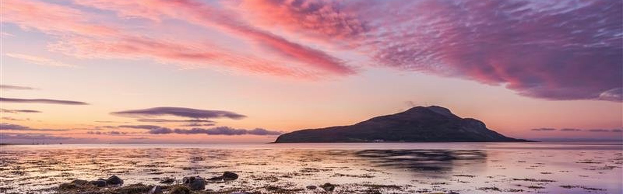 A view of Holy Isle from Lamlash Bay, Scotland. It is dusk and the sky is a beautiful mix of blue and purply pink clouds. The colours are reflected in shallow rock pools in the foreground.