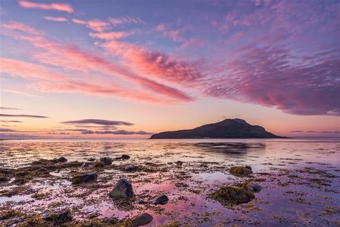 A view of Holy Isle from Lamlash Bay, Scotland. It is dusk and the sky is a beautiful mix of blue and purply pink clouds. The colours are reflected in shallow rock pools in the foreground.