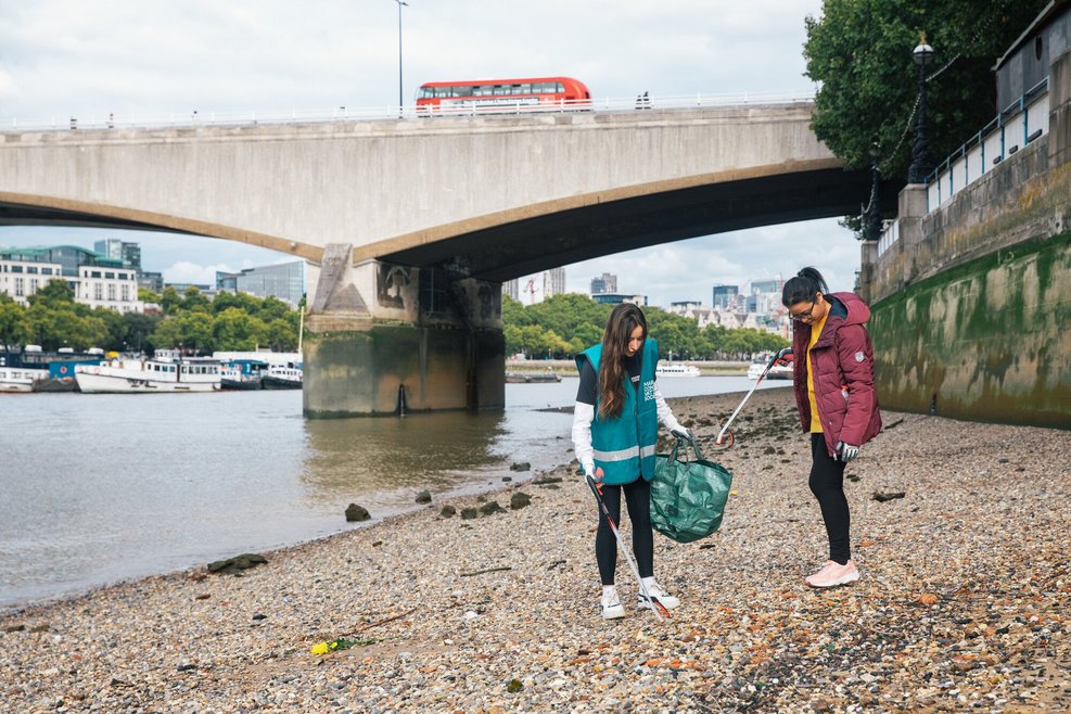 2 women picking up litter along an embankment. One is wearing a Marine Conservation Society safety vest and is holding a litter bag and litter picker.