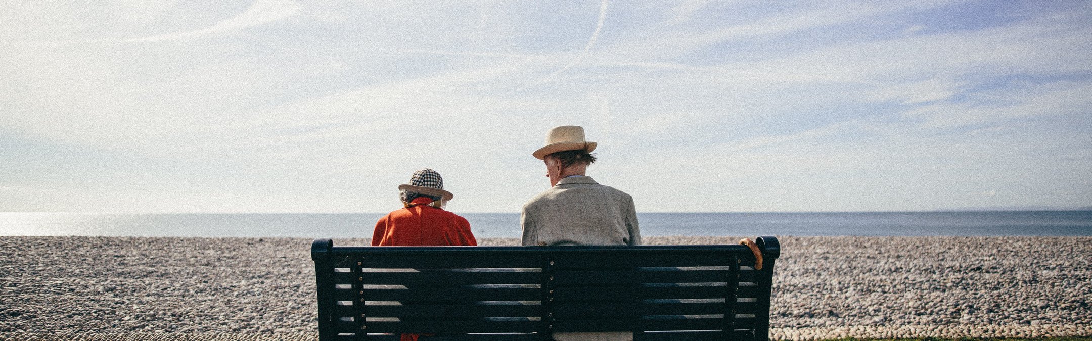 An elderly couple sitting on a dark green bench wth their backs to the camera. They are looking over a shingle beach toward a calm, blue sea. The man has a light coloured blazer and straw hat and the woman wears a red jacket and patterned bucket hat.