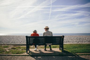 An elderly couple sitting on a dark green bench wth their backs to the camera. They are looking over a shingle beach toward a calm, blue sea. The man has a light coloured blazer and straw hat and the woman wears a red jacket and patterned bucket hat.