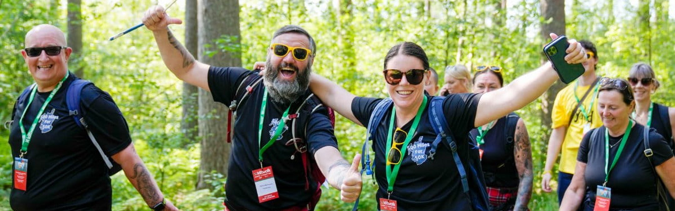 A group of smiling people wearing traditional red tartan kilts and black T-shirts hiking through a sunny, wooded area.