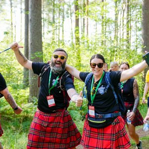 A group of smiling people wearing traditional red tartan kilts and black T-shirts hiking through a sunny, wooded area.