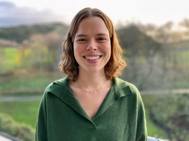 Trustee polly Burns smiles against a green landcaspe. She has short wavy brown hair and is wearing a moss-green jumper.