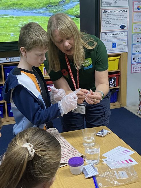 Sue Burton stands next to a schoolchild in a classroom. She is showing them a vial of water. On the table in front of them are beakers of water and water quality checking equipment