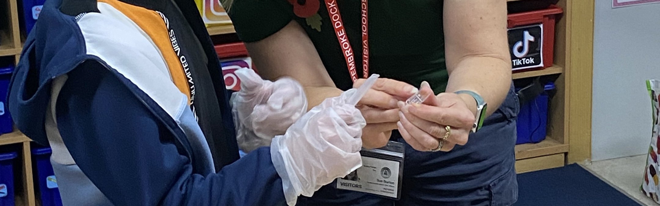 Sue Burton stands next to a schoolchild in a classroom. She is showing them a vial of water. On the table in front of them are beakers of water and water quality checking equipment