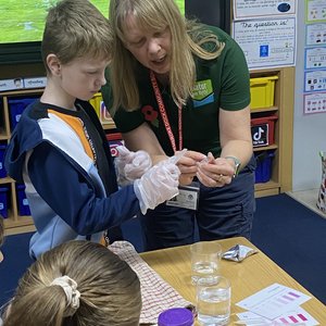 Sue Burton stands next to a schoolchild in a classroom. She is showing them a vial of water. On the table in front of them are beakers of water and water quality checking equipment
