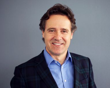 Trustee Stefano smiles against a plain grey background. It is a headshot style photo and he is wearing a blue shirt and green and blue checked jacket.