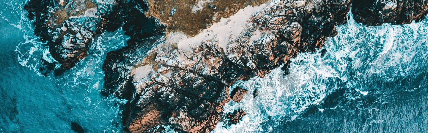 An aerial view of a rugged, rocky coastline where turquoise waves are crashing against the shore.
