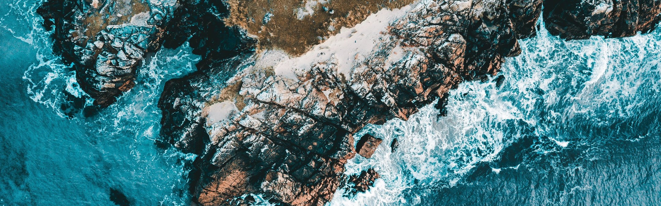 An aerial view of a rugged, rocky coastline where turquoise waves are crashing against the shore.