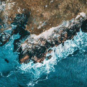 An aerial view of a rugged, rocky coastline where turquoise waves are crashing against the shore.
