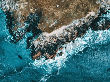 An aerial view of a rugged, rocky coastline where turquoise waves are crashing against the shore.