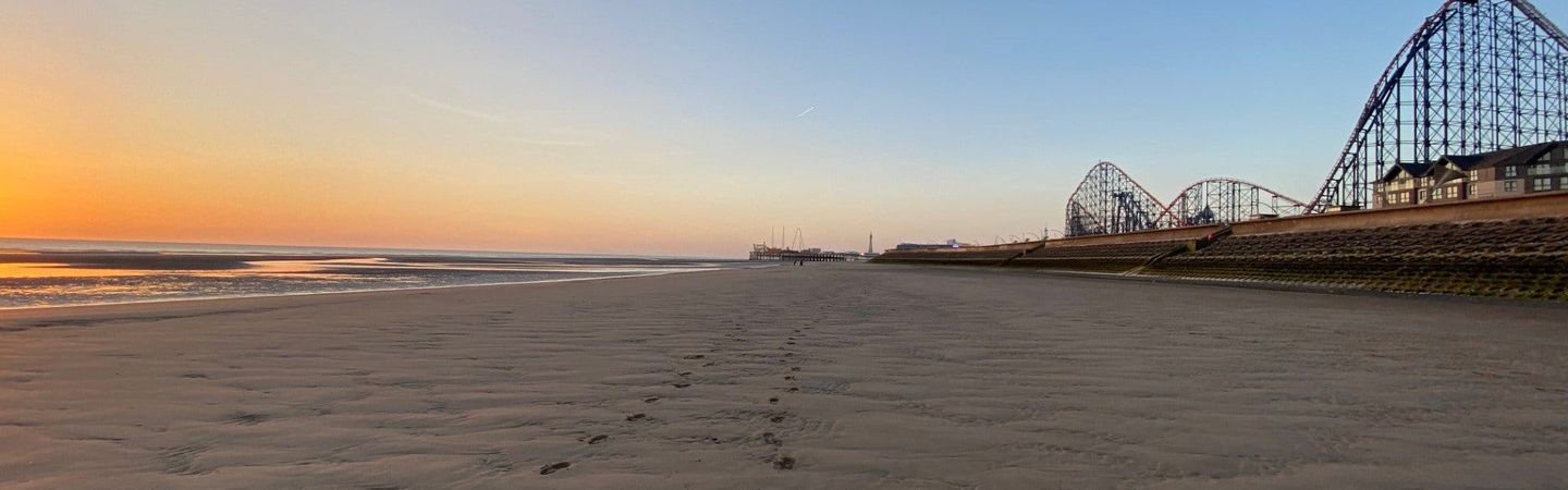 A beach with footprinds in the sand. A rollercoaster is in the background as part of a pleasure park