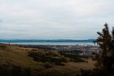 A wide-angle, slightly elevated view overlooking the city of Edinburgh, Scotland, under a grey, overcast sky. In the foreground, rolling green and brown hills slope downward.