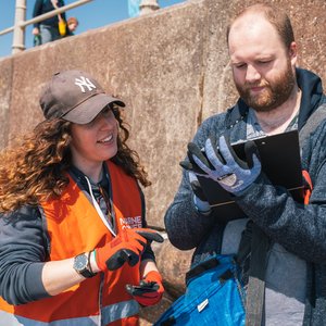 A woman with long, curly brown hair wearing a brown cap, orange high-vis vest and orange work gloves points toward a clipboard held by a man. The man is wearing a blue hoodie and blue work gloves. They are standing outdoors in front of a concrete wall.
