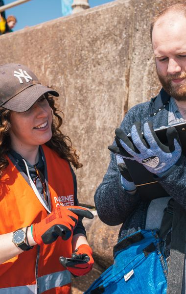 A woman with long, curly brown hair wearing a brown cap, orange high-vis vest and orange work gloves points toward a clipboard held by a man. The man is wearing a blue hoodie and blue work gloves. They are standing outdoors in front of a concrete wall.