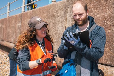 A woman with long, curly brown hair wearing a brown cap, orange high-vis vest and orange work gloves points toward a clipboard held by a man. The man is wearing a blue hoodie and blue work gloves. They are standing outdoors in front of a concrete wall.