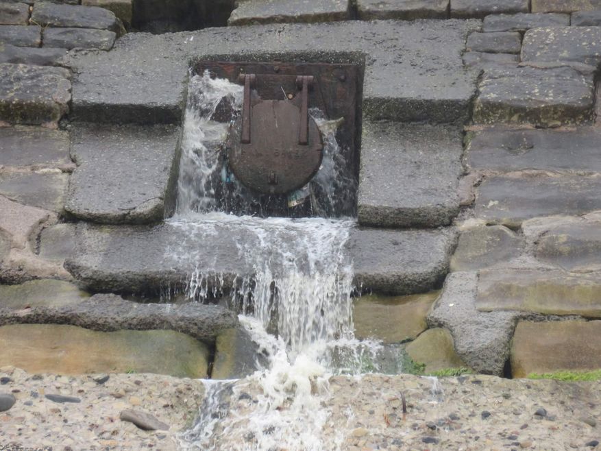Dirty water is flowing from a rusty metal flap gate in a concrete and stone wall, it is splashing onto the beach below