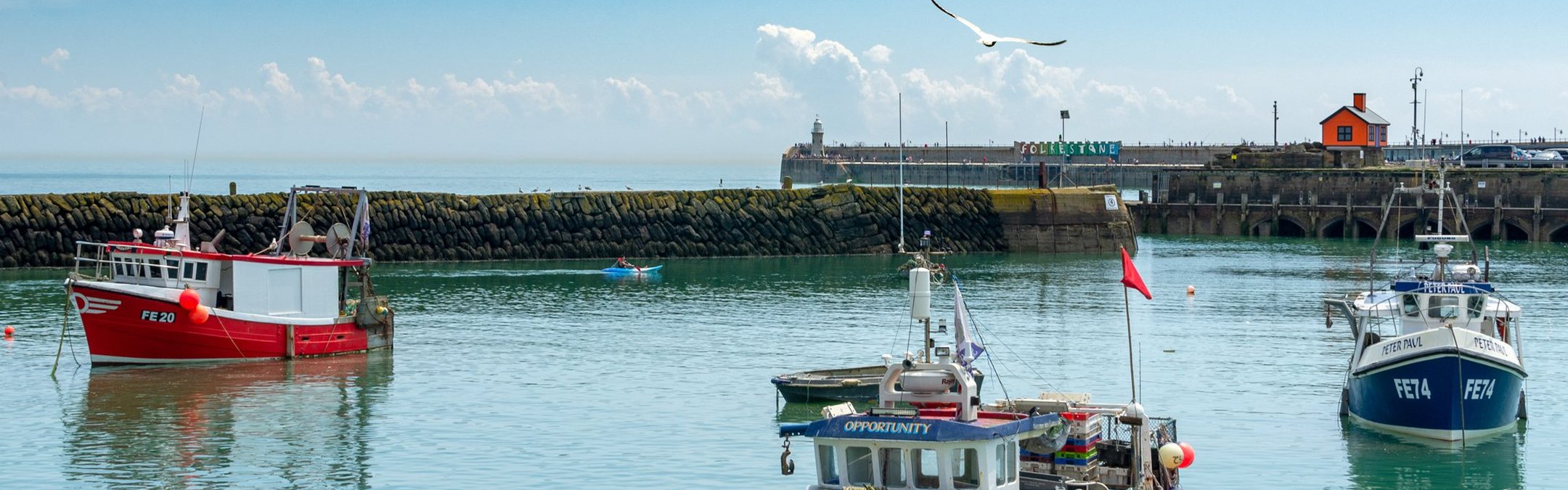 Several small, colourful fishing boats in Folkestone Harbour, with blue skies and the sun shining in the sky.