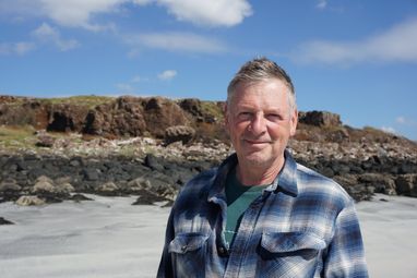 Trustee Mark Eckstein smiles on a sandy beach with a rocky bank in the background and a blue, slightly cloudy sky. He is wearing a checked shirt.