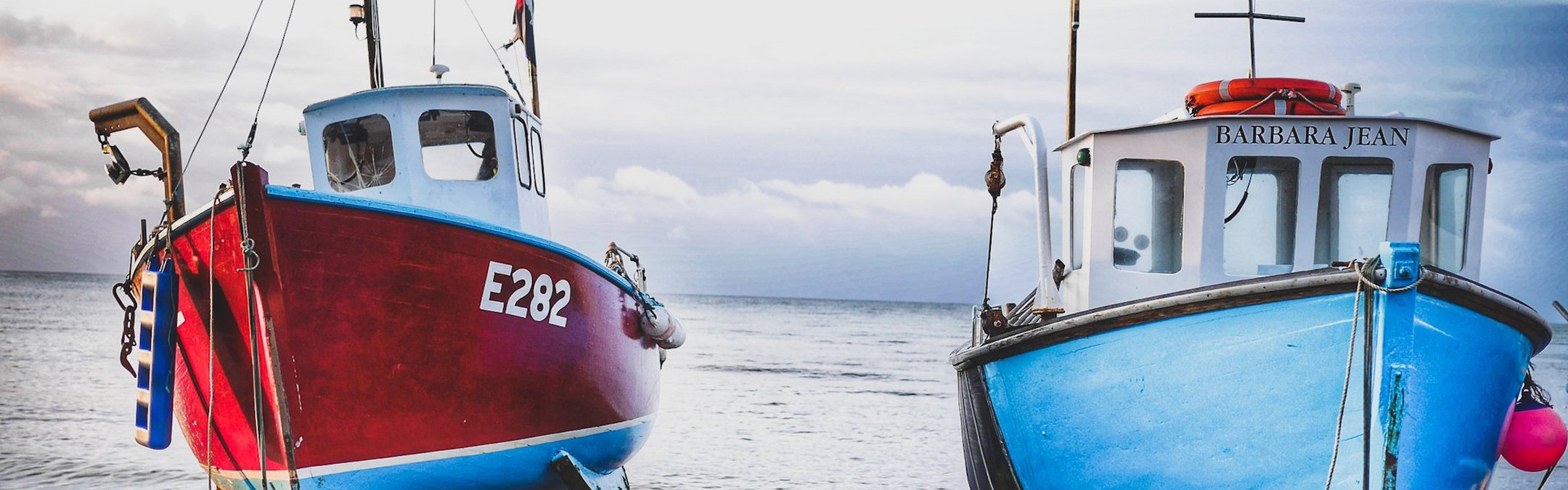 A close up of two fishing boats resting on pebbles. One boat is a vivid red colour, and the other is a bright blue colour.