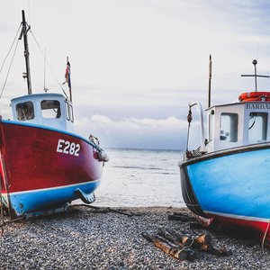 A close up of two fishing boats resting on pebbles. One boat is a vivid red colour, and the other is a bright blue colour.
