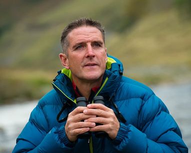 Iolo Williams stands against a Welsh mountain backdrop wearing a blue puffer coat. He is holding binoculars.