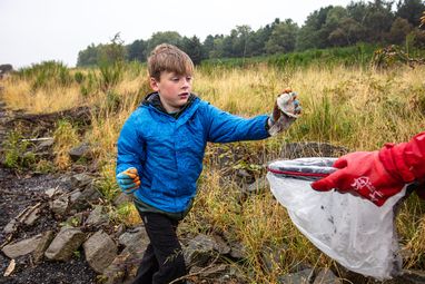A young boy in a blue jacket and red gloves using a litter picker to collect rubbish. He is putting a soiled wet wipe into a white bag which is being held by someone off camera.