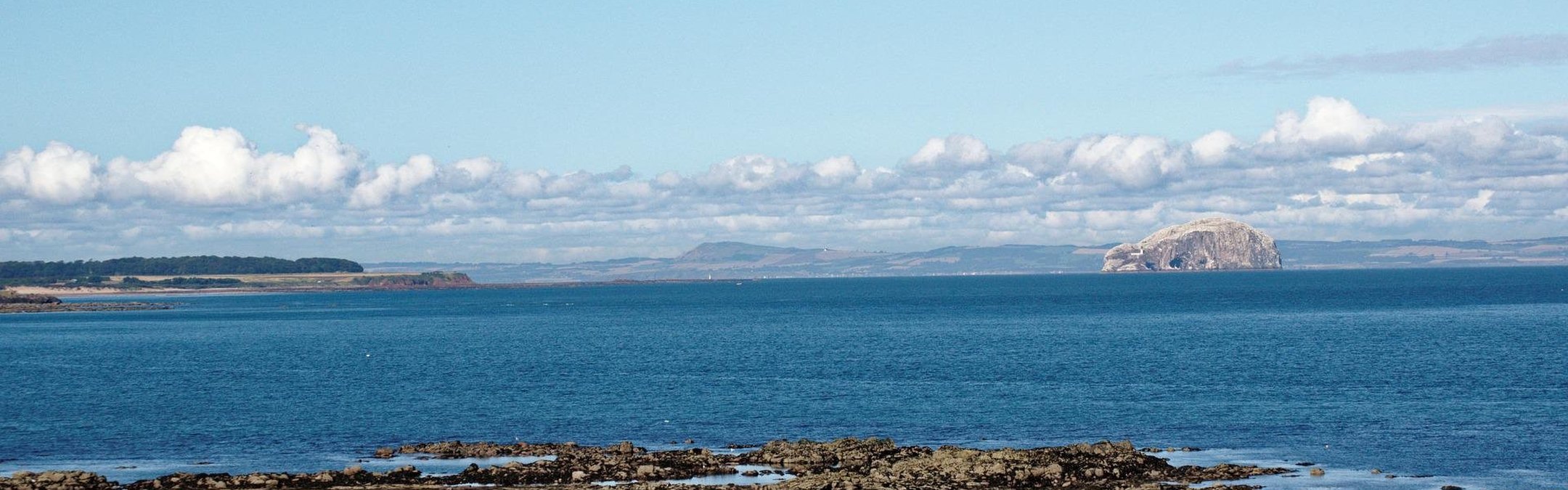 A view out over the Firth of Forth with a rocky shore in the foreground and a beautiful view across the water. The sky is blue and the clouds sit low in the sky.