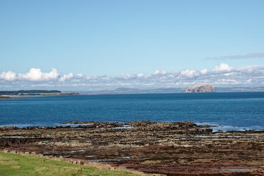 A view out over the Firth of Forth with a rocky shore in the foreground and a beautiful view across the water. The sky is blue and the clouds sit low in the sky.