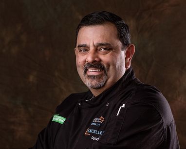 Chef Cyrus Todiwala smiles at the camera wearing chef's blacks against a black background.