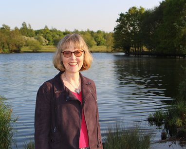 A picture of a smiling Amanda Nobbs, the Marine Conservation Society's Chair. She is standing in front of a calm lake backed with trees and has short blonde hair, a brown jacket and pink shirt.