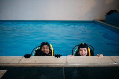 2 children learning to dive at Dive Project Cornwall. They are in a pool wearing diving gear and smiling at the camera