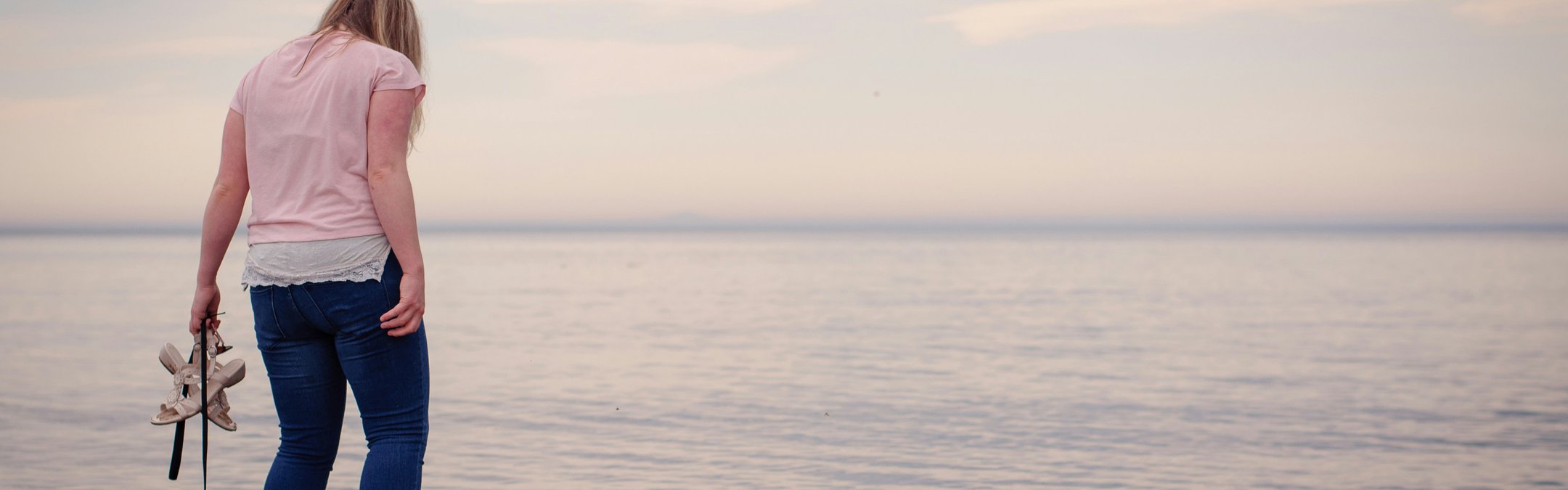 a person standing at the edge of the sea while holding a pair of shoes