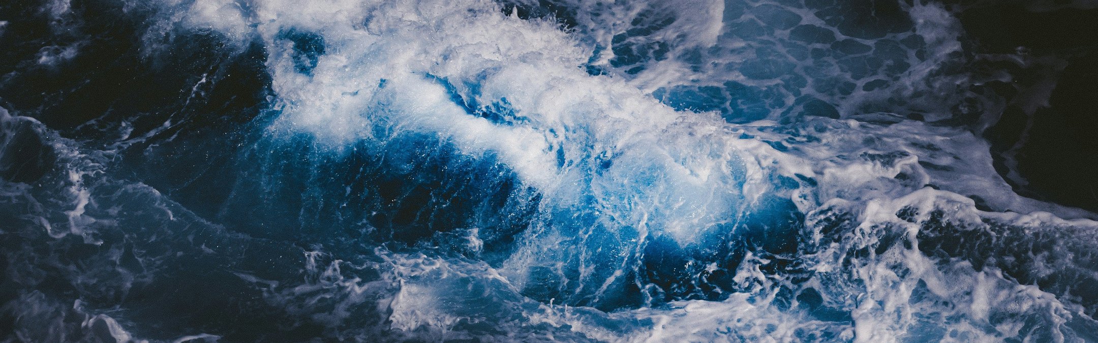 An aerial view of dark ocean waves. The water is indigo blue and white peaks of crashing waves can be seen.