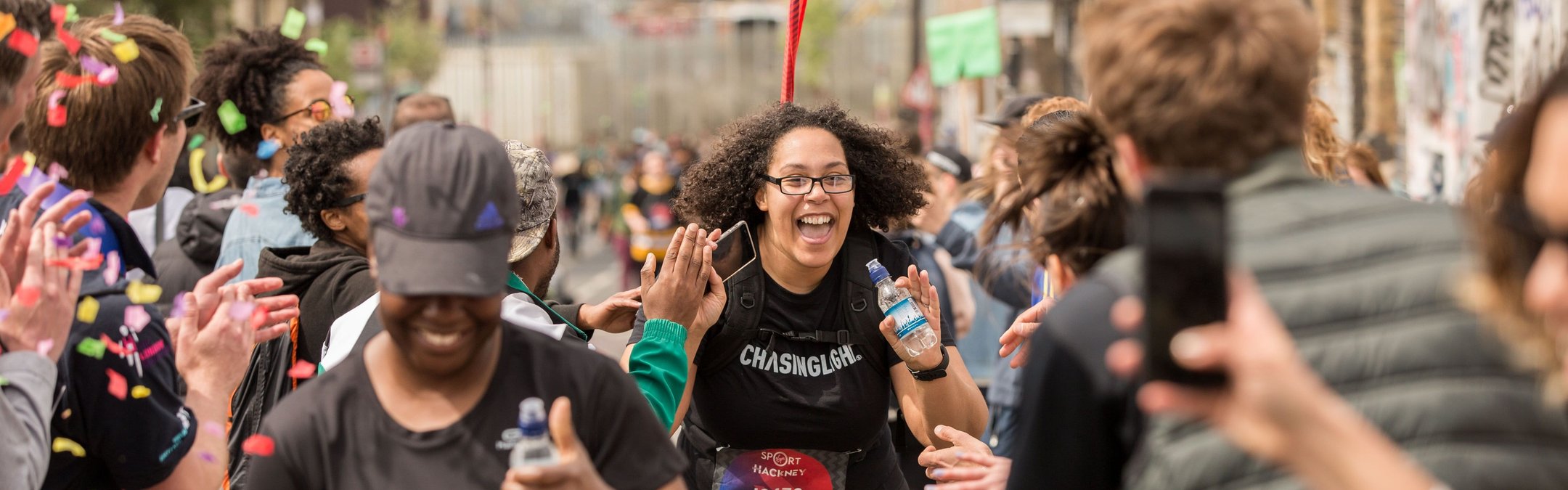 A happy runner with curly hair and glasses, wearing a black t-shirt, celebrates as she approaches a finish line. She is surrounded by supporters
