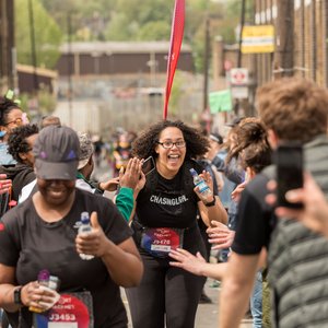 A happy runner with curly hair and glasses, wearing a black t-shirt, celebrates as she approaches a finish line. She is surrounded by supporters