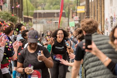 A happy runner with curly hair and glasses, wearing a black t-shirt, celebrates as she approaches a finish line. She is surrounded by supporters