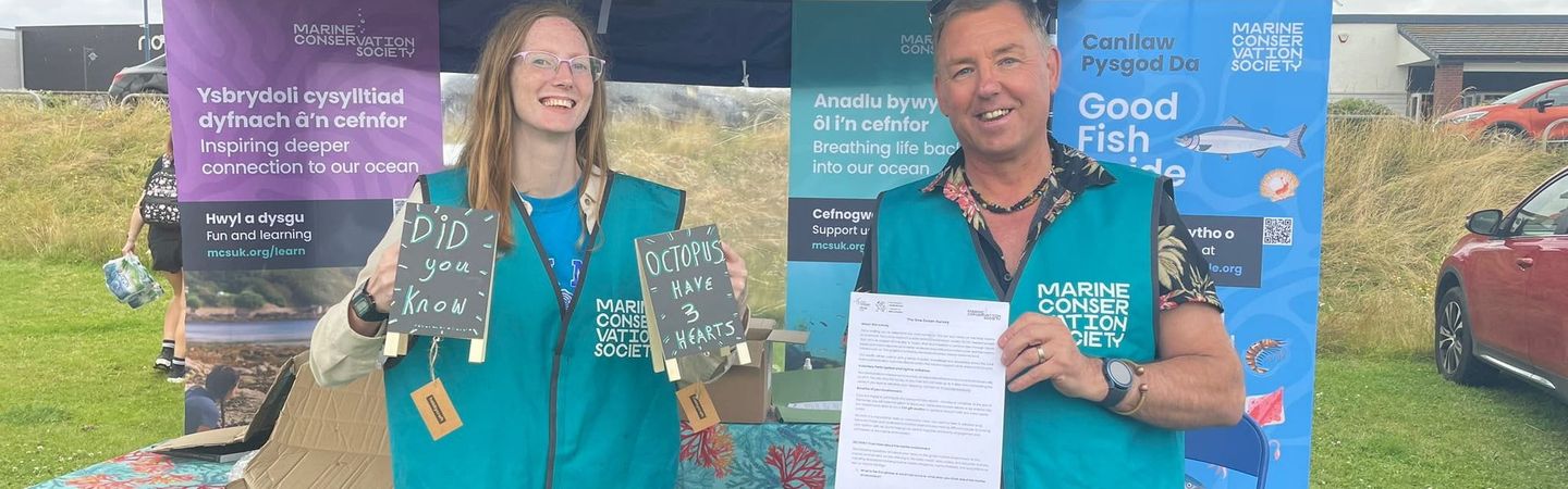 A man and a woman in turquoise vests stand behind a table at a Marine Conservation Society event, holding signs and a document. Banners with Welsh and English text are visible in the background.