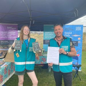 A man and a woman in turquoise vests stand behind a table at a Marine Conservation Society event, holding signs and a document. Banners with Welsh and English text are visible in the background.